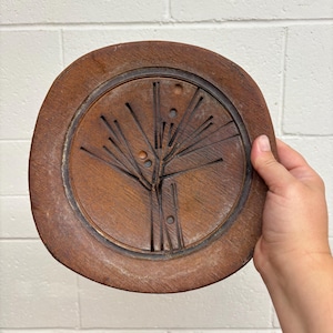 May include: A round, brown wooden plate featuring a carved, abstract sunburst or tree-like pattern of thin, straight lines radiating from a central point, accented by small, circular indentations within the circular, recessed centre area.