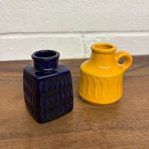 May include: A square-shaped dark blue ceramic vase with a textured vertical pattern and a round opening, paired with a rounded yellow ceramic pitcher featuring a side handle and a repeating embossed pattern around the base of the vessel.