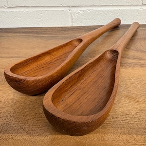 Op de afbeelding: Twee houten serveerlepels met lange handvatten en ovale kommen. De lepels zijn gemaakt van lichtbruin hout en hebben een gladde, gepolijste afwerking. De lepels worden gepresenteerd op een houten oppervlak.