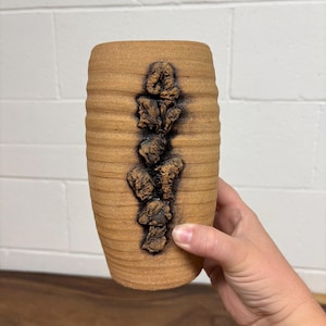 May include: A cylindrical tan ceramic vase with a ribbed, textured surface. A vertical column of dark, rough, organic-shaped clay protrusions runs down the centre of the vase, creating a stark contrast against the smooth, matte tan body.