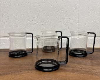 Op de afbeelding: Vier heldere glazen mokken met zwarte metalen handvatten en bodems staan op een houten oppervlak. De mokken zijn cilindrisch en leeg. De achtergrond is een witte bakstenen muur.