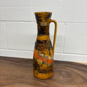 Puede incluir: Un jarrón de cerámica Carstens West Germany vintage. El jarrón presenta un diseño texturizado en tonos marrón, naranja y amarillo. Tiene un asa y una etiqueta que dice "Carstens". El jarrón está sobre una superficie de madera.