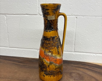May include: A vintage Carstens West Germany ceramic vase. The vase features a textured, layered design in shades of brown, orange, and yellow. It has a handle and a label that reads "Carstens". The vase is on a wooden surface.