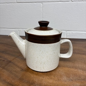Puede incluir: Una tetera de cerámica con un cuerpo color crema moteado y una banda marrón oscuro alrededor de la parte superior. La tapa y el asa también son de color marrón oscuro. La tetera tiene un pico y un asa.