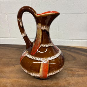 May include: A brown ceramic jug with a curved handle and a flared spout. The jug has a glossy finish with a design of orange, white, and tan vertical stripes. The interior of the spout is orange.