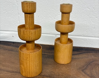 Puede incluir: Dos candeleros de madera de color marrón claro. Cada uno tiene una base cilíndrica, una sección media en forma de cuenco y una parte superior diseñada para una vela. Los candeleros se exhiben sobre una superficie de madera.