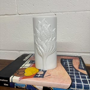 May include: White ceramic vase with a raised design of stylized leaves. The vase is cylindrical and has a smooth, glossy finish.