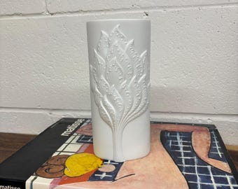 May include: White ceramic vase with a raised design of stylized leaves. The vase is cylindrical and has a smooth, glossy finish.