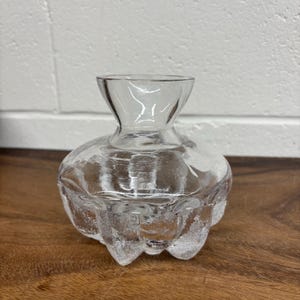 May include: A clear glass bud vase with a rounded base and a slender neck. The vase has a textured, possibly frosted, appearance. The base has a unique, petal-like design. The vase is on a wooden surface.