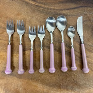 May include: A set of eight stainless steel utensils featuring light purple, iridescent, cylindrical handles with rounded ends. The collection includes four forks, two large spoons, one small spoon, and one flat-edged butter knife.