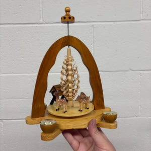 Pode incluir: Uma pirâmide de Natal de madeira com uma árvore central, figuras de veados e castiçais. A pirâmide tem um design giratório e é feita de madeira castanha clara e escura. O design inclui um arco decorativo e uma pequena casa.