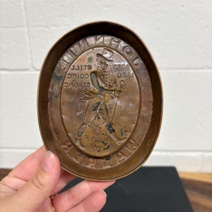 Op de afbeelding: Ovale koperkleurige metalen schaal met reli&euml;ftekst en een figuur van een man met hoge hoed. De tekst bevat de woorden "Born 1850" en "Johnnie Walker". De schaal heeft een opstaande rand.