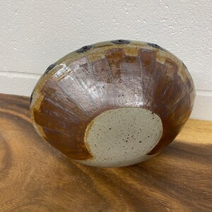 Puede incluir: Un cuenco de cerámica marrón y blanco con una superficie texturizada y un esmalte marrón. El cuenco tiene una forma redonda y un borde ancho. Hay pequeños agujeros alrededor del borde del cuenco.