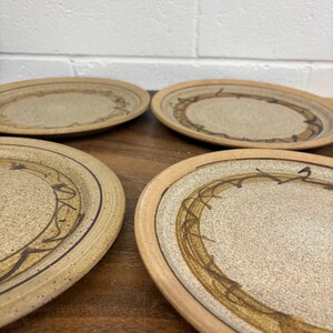 May include: Four beige ceramic plates with a speckled surface and a brown decorative border. Each plate has a unique brown design in the centre. The plates are arranged on a wooden surface.
