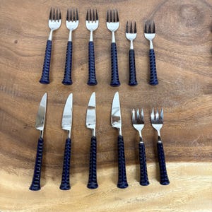 May include: A collection of stainless steel forks and knives with navy blue handles. The forks have four prongs, and the knives have a smooth blade. The handles have a textured, ridged design. The set is arranged on a wooden surface.