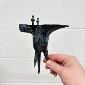 May include: A small, dark green bronze sculpture of a Chinese ceremonial wine cup, known as a "gui", with intricate carvings and three legs.