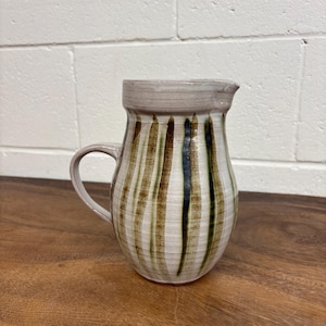 May include: A ceramic pitcher with a light beige base and vertical stripes in shades of brown and dark green. The pitcher features a rounded body, a curved handle on the left side, and a small pouring spout at the top rim with a textured finish.