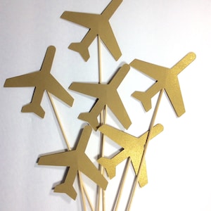May include: Six gold paper airplanes on wooden sticks. The airplanes are arranged in a cluster, with the sticks pointing upwards.
