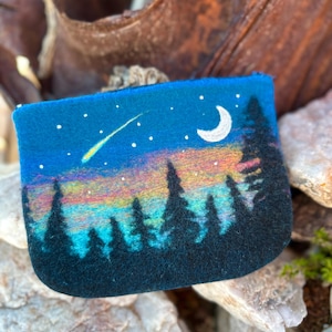 May include: A blue felt pouch with a night sky design. The design features a crescent moon, stars, and a shooting star over a colorful sunset and a silhouette of pine trees. The pouch has a zipper closure.
