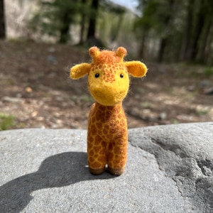 May include: A small, hand-crafted felt giraffe toy with a yellow body and brown spots. The giraffe has a friendly face with black eyes and a small, pink nose.
