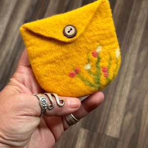 May include: A small yellow felt pouch with a button closure and a floral design. The pouch is held in a hand with a ring on the finger.