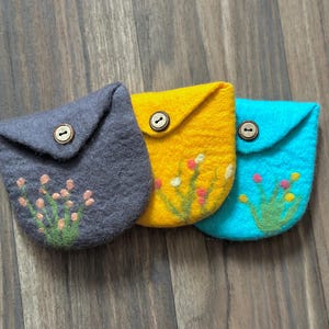 May include: Three felt pouches in grey, yellow, and turquoise, each with a button closure. The pouches are decorated with embroidered floral designs in pink, green, and white. The pouches are approximately 10 cm wide.