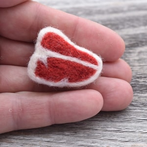 May include: A small, red felt sculpture of a steak, with a white felt outline. The steak is held in a person's hand.