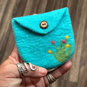 May include: A small turquoise felt pouch with a brown button closure and a green and yellow embroidered flower design.