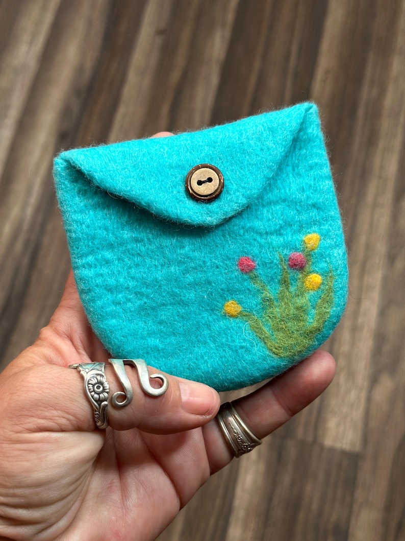 May include: A small turquoise felt pouch with a brown button closure and a green and yellow embroidered flower design.