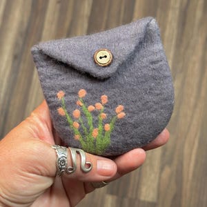 May include: A small gray felt pouch with a flap closure and a wooden button. The pouch has a floral design embroidered on the front in shades of orange and green.