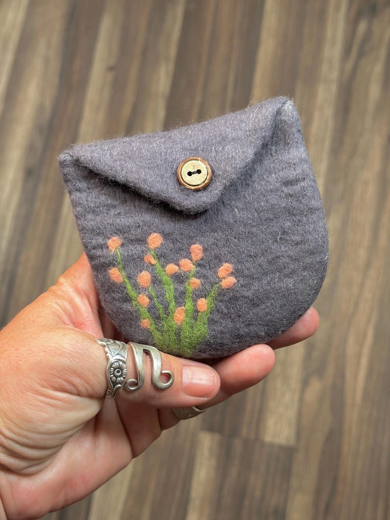 May include: A small gray felt pouch with a flap closure and a wooden button. The pouch has a floral design embroidered on the front in shades of orange and green.