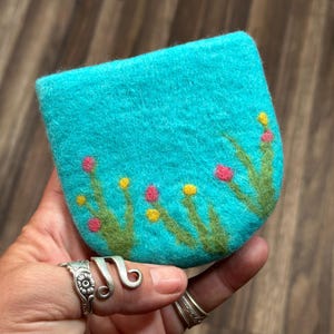 May include: A teal blue felt coin purse with a hand-stitched design of green grass and pink and yellow flowers.