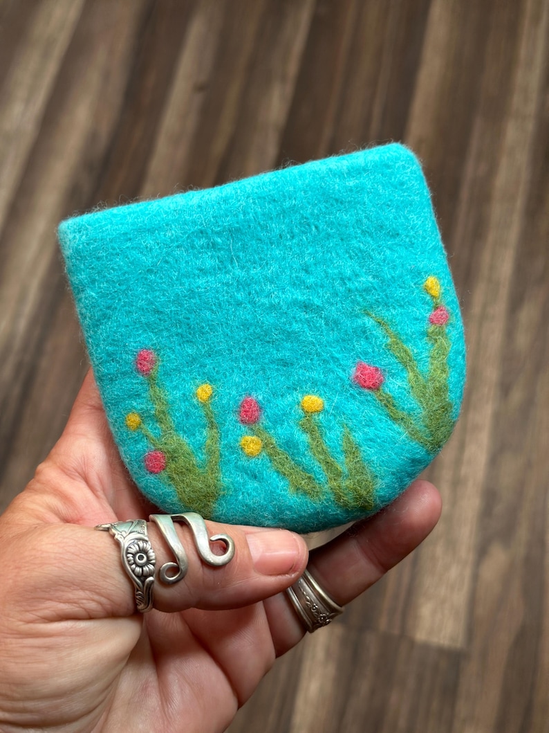May include: A teal blue felt coin purse with a hand-stitched design of green grass and pink and yellow flowers.