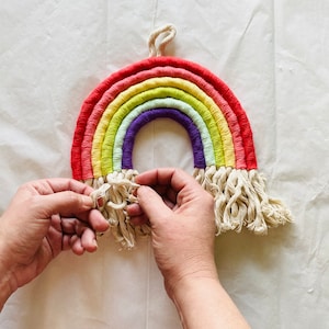 May include: A rainbow-colored macrame wall hanging with a white fringe. The hanging is made of yarn and is in the shape of a rainbow.