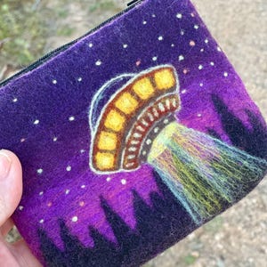 May include: A purple felt pouch with a zipper closure. The pouch features a colorful illustration of a UFO emitting a beam of light over a silhouetted landscape. White dots represent stars in the night sky.