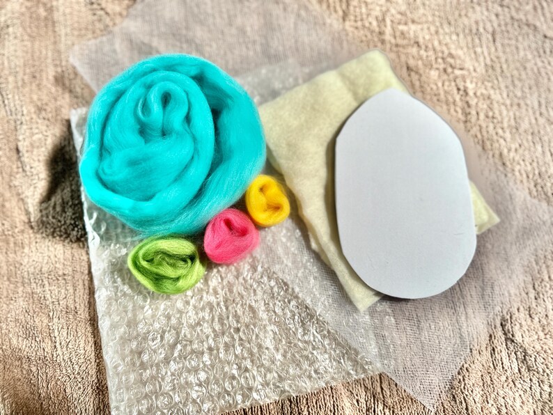 May include: A set of wool roving for needle felting, including a light blue, pink, yellow, and green roving. The set also includes a white felt pad and a clear plastic sheet.