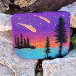 May include: A felted pouch featuring a vibrant sunset scene with silhouetted trees and two shooting stars against a purple starry sky. The pouch has a zipper closure and is made of felt.