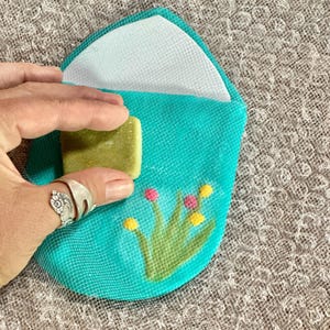 May include: A teal blue fabric pouch with a white lining and a floral design. The pouch is open and a green bar of soap is inside.
