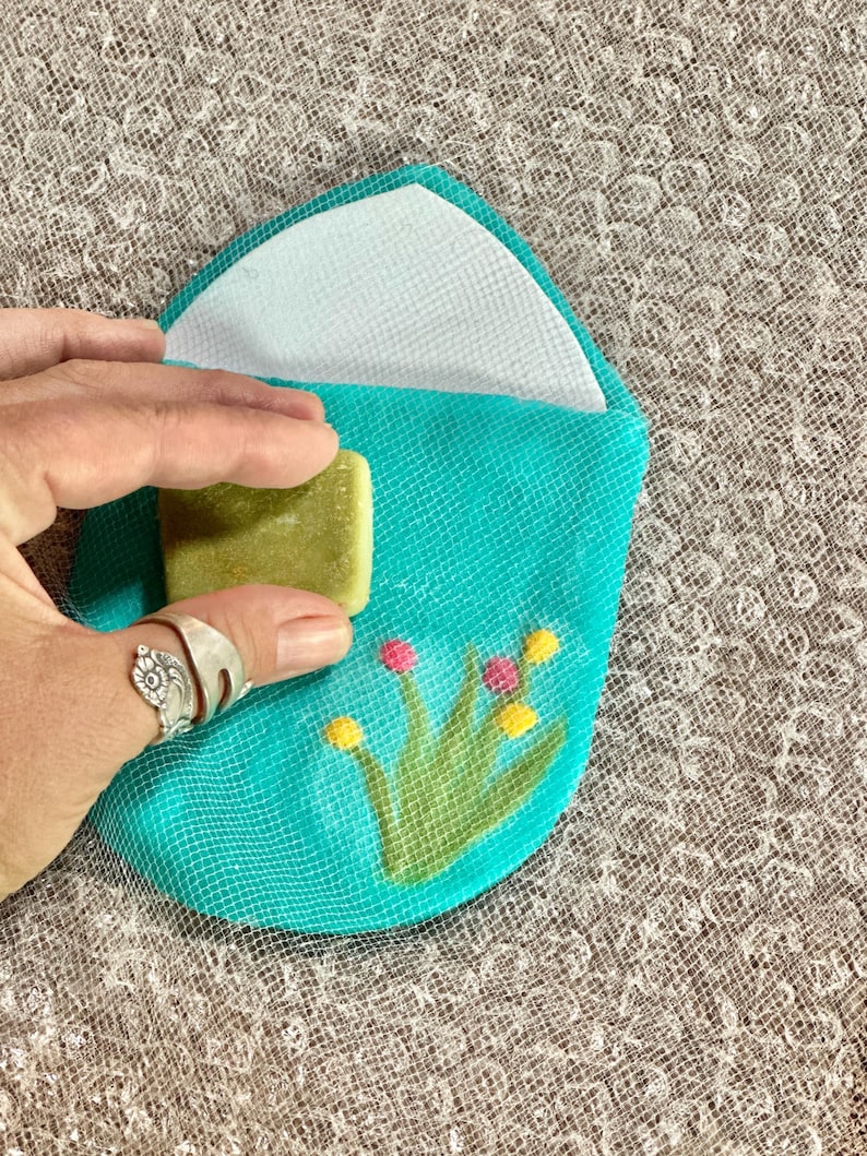 May include: A teal blue fabric pouch with a white lining and a floral design. The pouch is open and a green bar of soap is inside.
