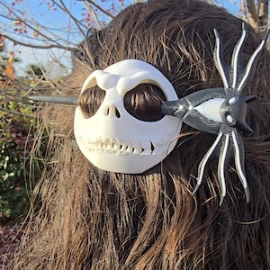 May include: A white skull-shaped hair clip with a black and white bat-like design. The hair accessory is a unique, whimsical item, perfect for Halloween or cosplay. The clip is set against a backdrop of brown hair and a blue sky.