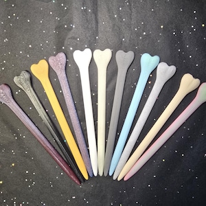 May include: A collection of heart-topped hair sticks in various colors, including black, yellow, purple, white, gray, and blue. The sticks are arranged in a fan shape against a black background with glitter.