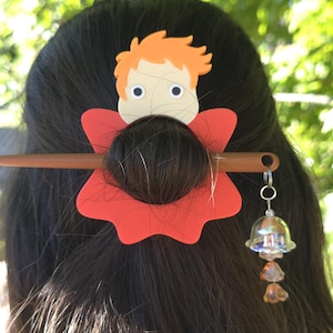 Op de afbeelding: Een haaraccessoire met een platte cartoonfiguurkop met oranje haar op een rode, bloemvormige basis. Een bruine houten haarstok houdt het geheel vast, met een kleine, doorzichtige, iriserende belhanger die aan het uiteinde van de stok hangt.