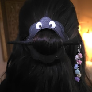 Może przedstawiać: Czarny dodatek do włosów o fantazyjnym designie, z ciemnoszarym kształtem chmury z białymi oczami. Czarny patyk utrzymuje włosy na miejscu. Srebrny łańcuszek z pastelowymi gwiazdkami wisi.