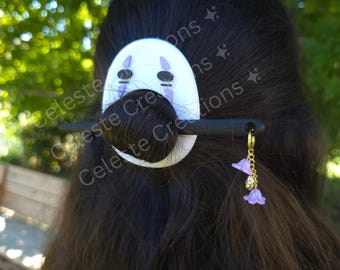 May include: A hair accessory featuring a white oval face plate with black and purple markings, secured by a black hair stick. A gold-toned chain hangs from the stick, adorned with two small purple bell-shaped charms and a clear crystal bead.