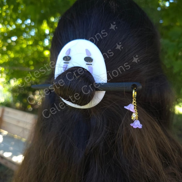 May include: A hair accessory featuring a white oval piece with a black and purple character face design, secured by a black hair stick. A gold-toned hoop charm with two small purple bell-shaped flowers and a clear crystal bead hangs from the stick.