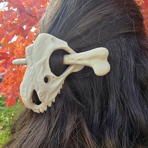 May include: A cream-colored hair barrette shaped like a dinosaur skull and bone, worn in dark brown hair. The barrette is detailed with eye sockets and teeth. The background features vibrant red and orange autumn leaves.