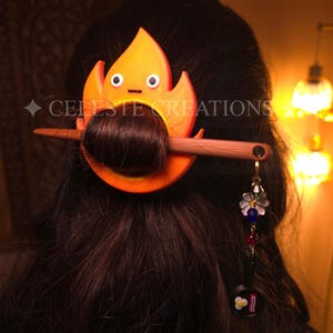 May include: A hair accessory featuring an orange flame-shaped character with two eyes and a small mouth, secured by a brown wooden hair stick. A dangling charm hangs from the stick, displaying a small frying pan with bacon and eggs.