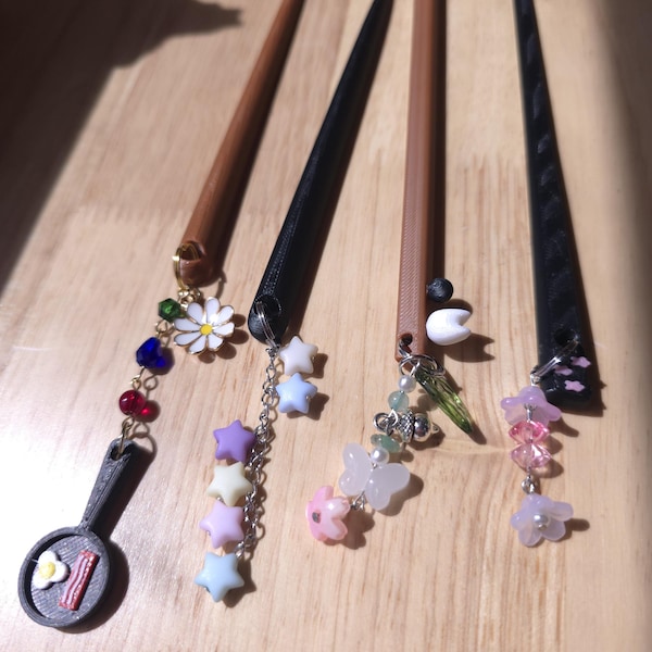 May include: Four decorative hair sticks with unique designs. One features a small frying pan charm with a fried egg and bacon, another has pastel star charms, a third has butterfly and bead charms, and the last has flower charms. The sticks are brown, black, and tan.
