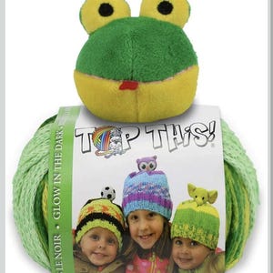 May include: A green and yellow frog plush toy rests on a skein of light green yarn. The yarn is wrapped around a package with the text "TOP THIS!" and images of children wearing knitted hats. The text "GLOW IN THE DARK" is also visible.