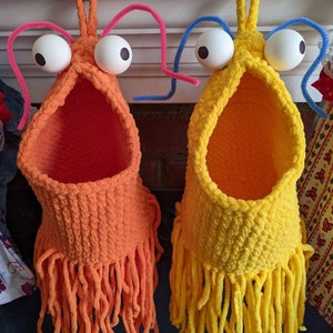 May include: Two handmade, whimsical crochet baskets, one orange and one yellow, each with large googly eyes and fringe. The orange basket has pink pipe cleaner antennae, while the yellow one has blue. These decorative baskets are perfect for storing small items.